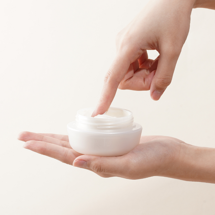 Hand holding a small white container of cream against a light background