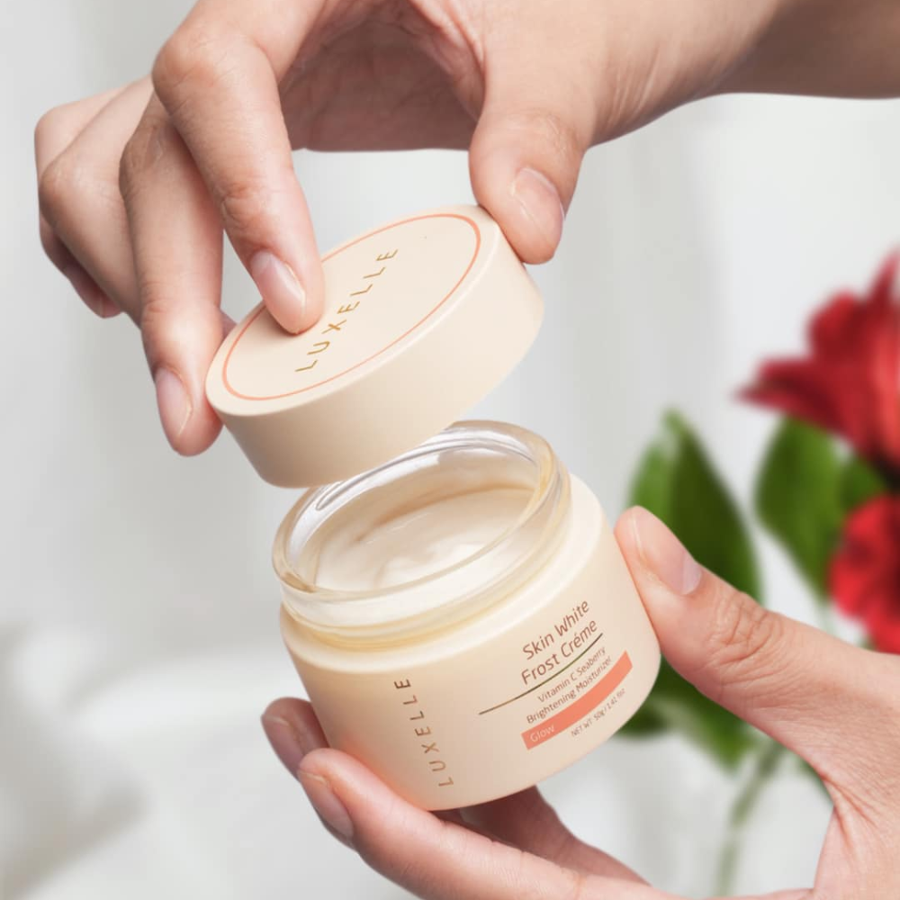 Person holding a jar of Luxelle Skin White Foot Creme with a blurred floral background
